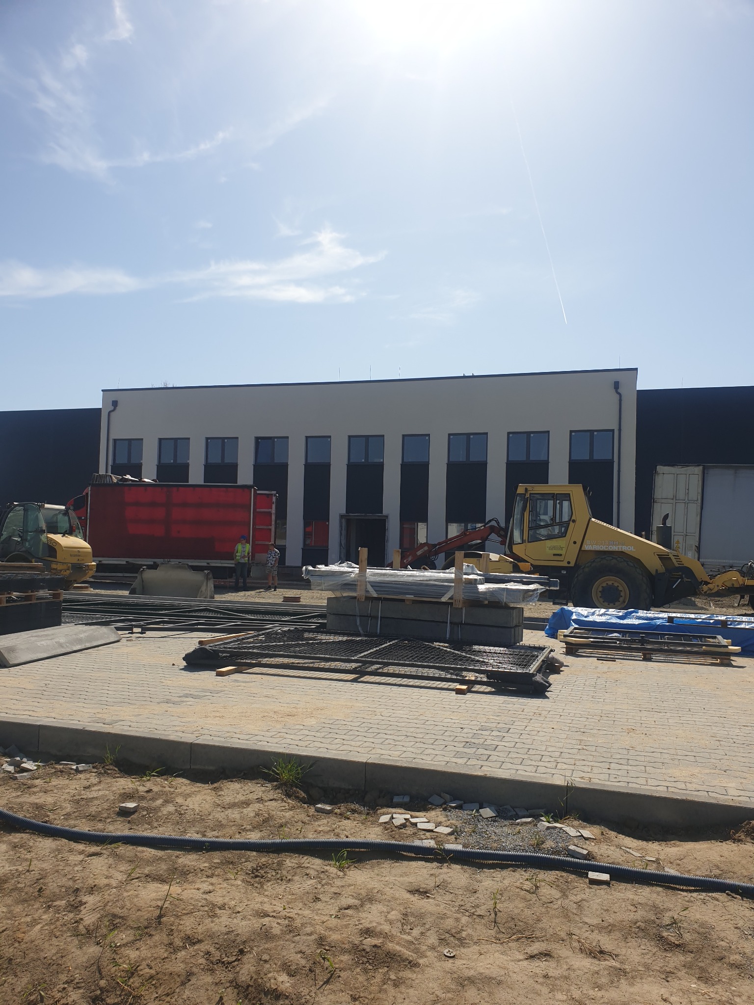 Plac budowy przed nowoczesnym budynkiem magazynowym z żółtą ładowarką kołową i czerwoną ciężarówką w słoneczny dzień.