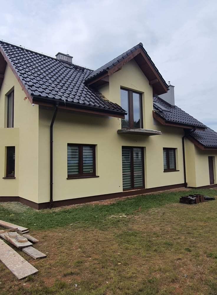 Żółta elewacja domu jednorodzinnego z ciemnobrązowymi oknami i dachem z dachówki ceramicznej, widoczne deski na trawniku.