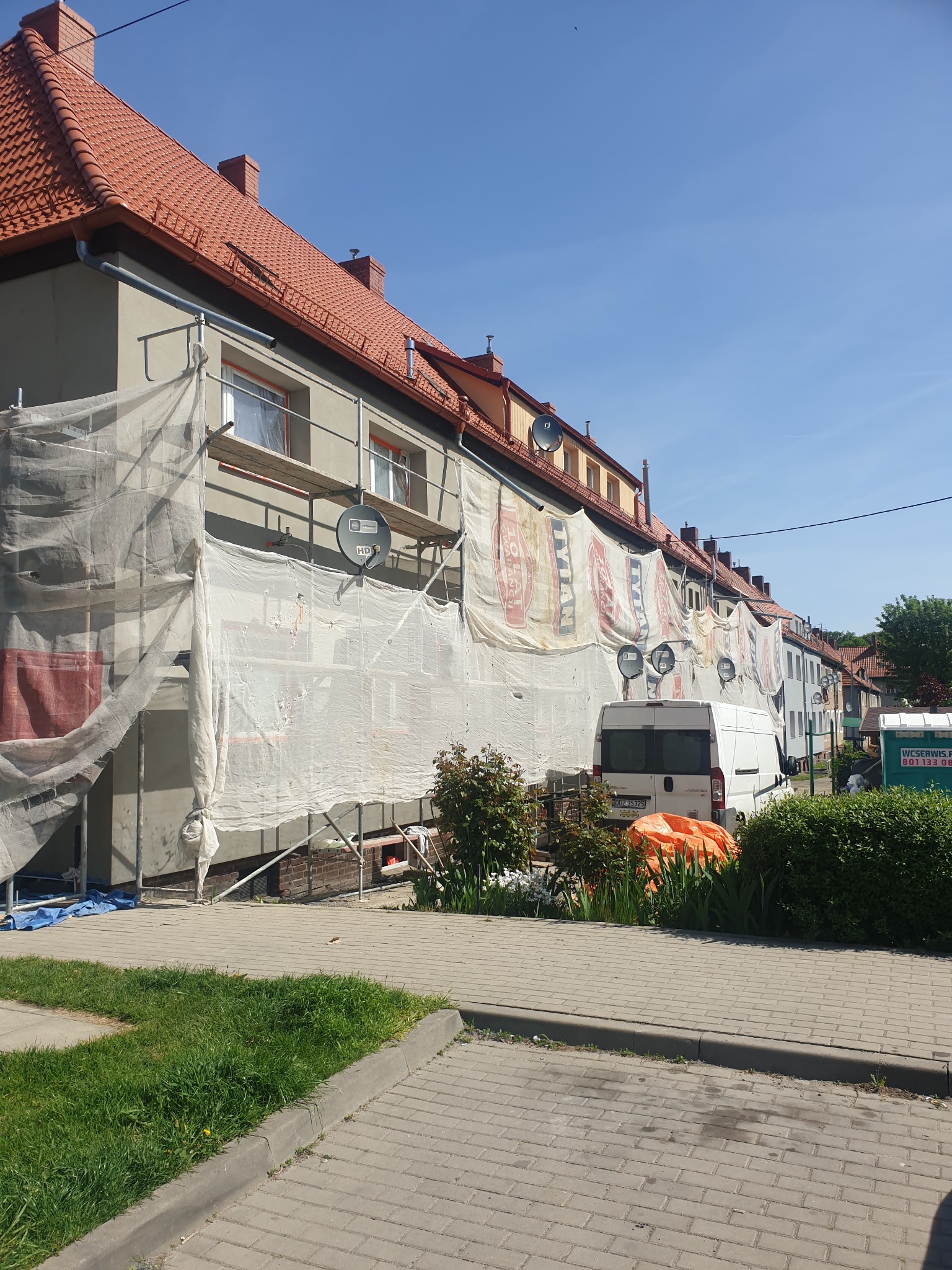 Ocieplanie elewacji szeregu domów jednorodzinnych, osłoniętych siatką budowlaną i rusztowaniami, z widocznym samochodem dostawczym i częściowo wykończoną elewacją.