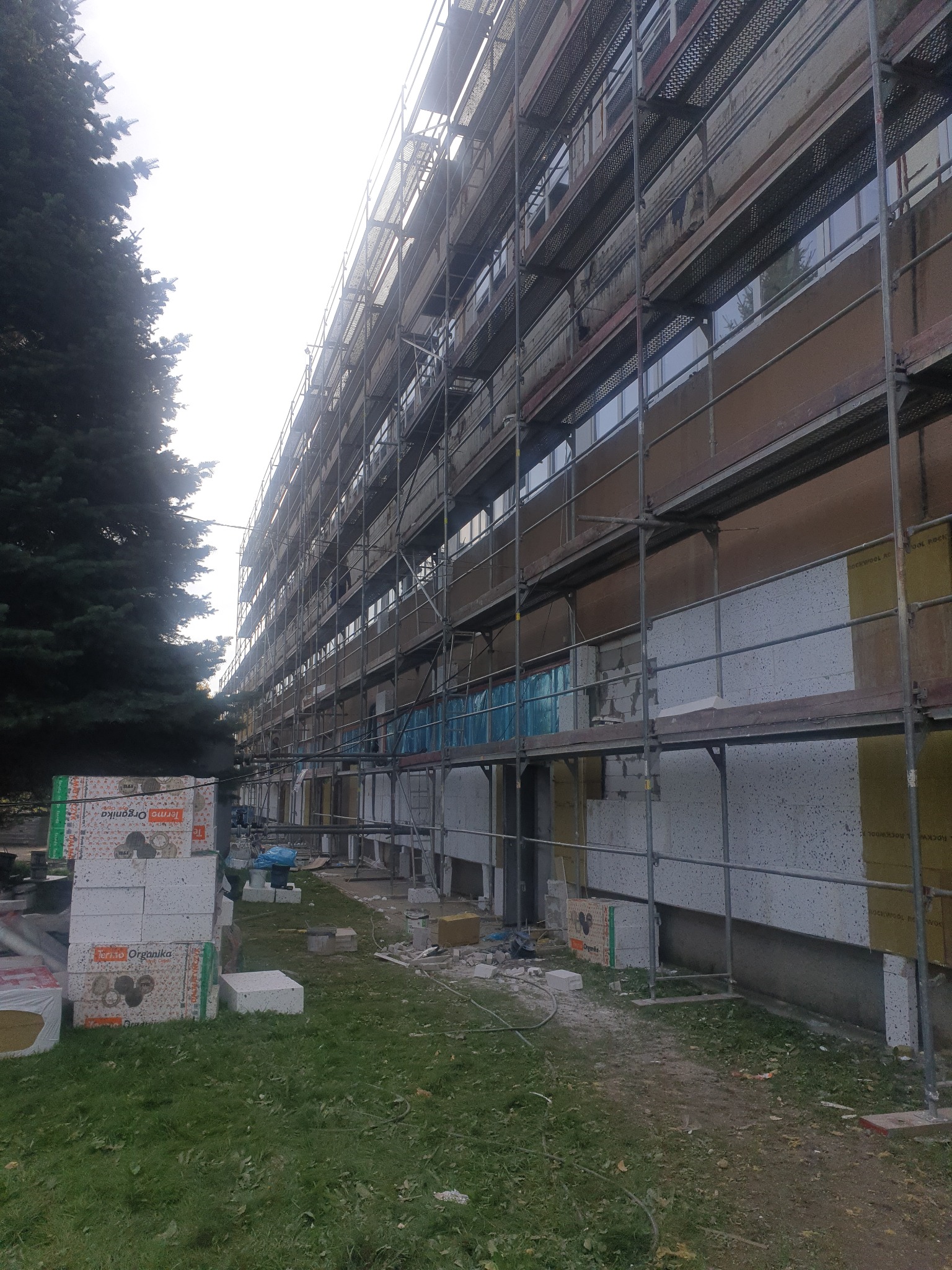 Budynek wielorodzinny w trakcie prac elewacyjnych, widoczne rusztowania, styropian i materiały budowlane na trawniku.