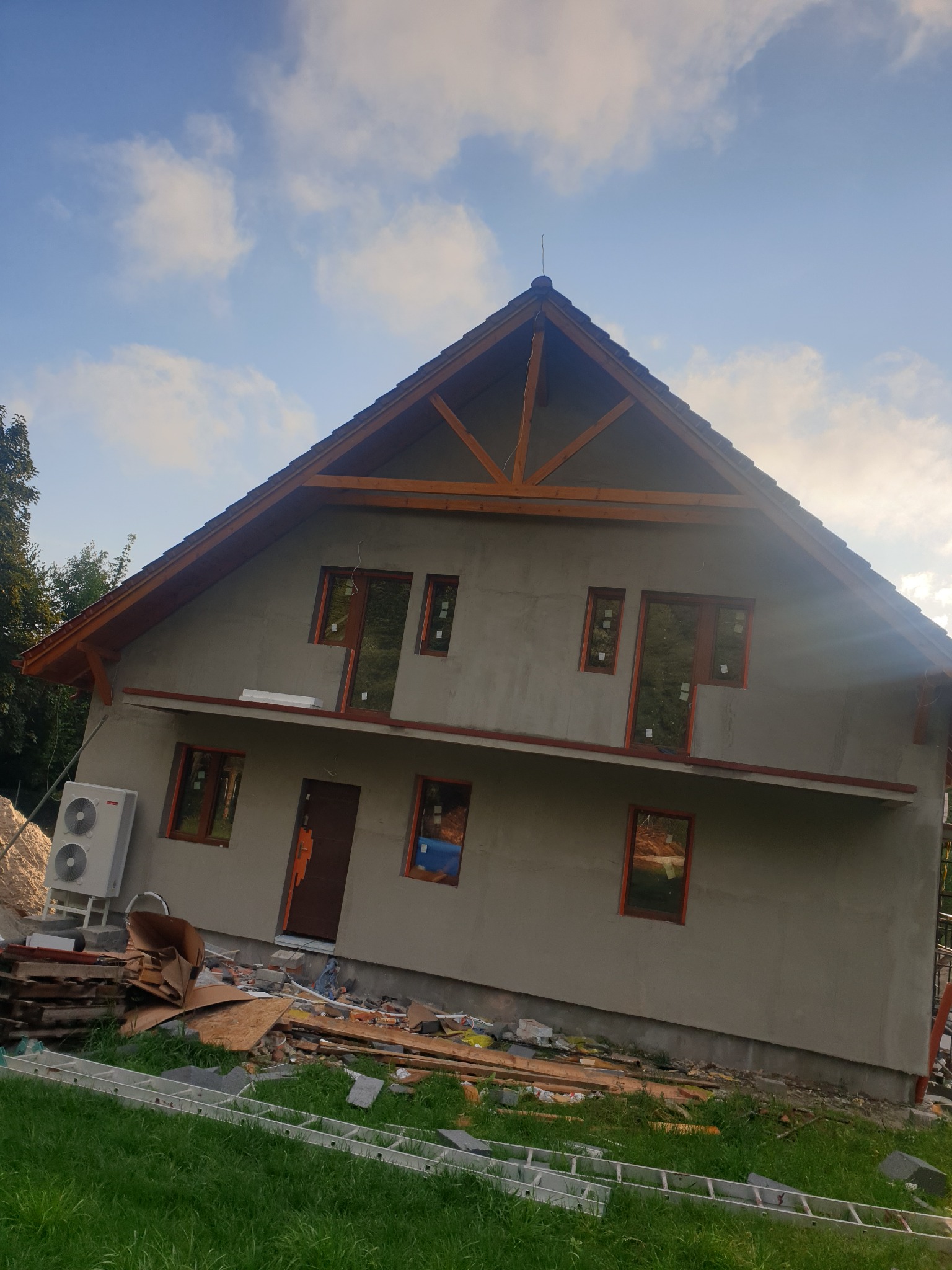 Surowy stan elewacji domu jednorodzinnego z widocznymi oknami w drewnianych ramach, jednostką klimatyzacji na zewnątrz i stertą materiałów budowlanych na trawniku.