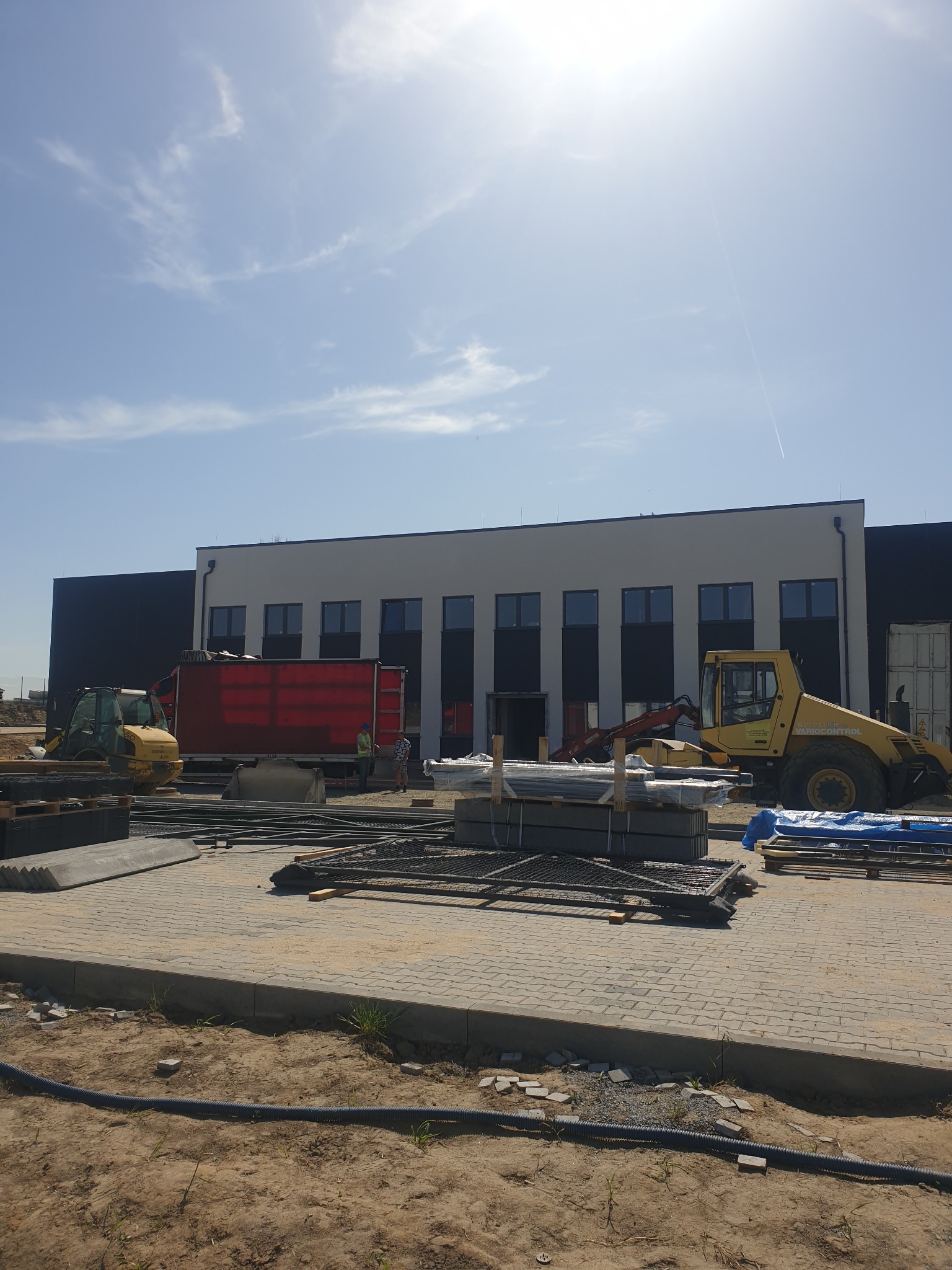 Plac budowy przed budynkiem z szarą elewacją i czarnymi elementami, widoczny żółty sprzęt budowlany i materiały przygotowane do prac elewacyjnych.