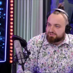 Osoba z brodą w koszuli w kwiaty, w słuchawkach, siedzi przed mikrofonem w studio nagraniowym w Rzeszowie, w tle oświetlenie LED i monitory.