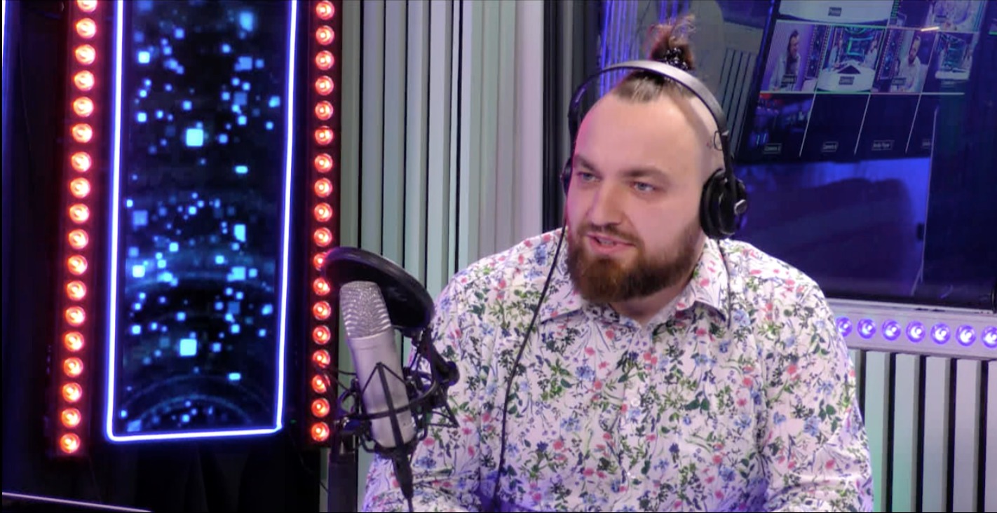 Osoba z brodą w koszuli w kwiaty, w słuchawkach, siedzi przed mikrofonem w studio nagraniowym w Rzeszowie, w tle oświetlenie LED i monitory.