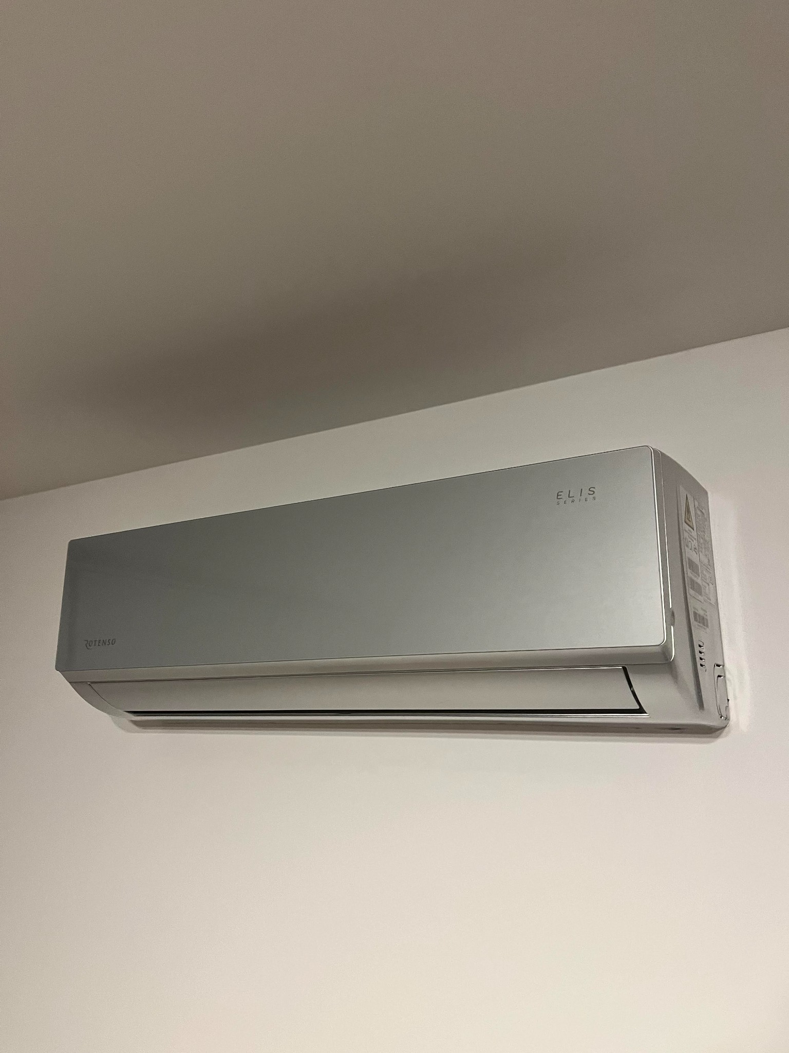 Nowoczesna, srebrna klimatyzacja marki Rotenso Elis Series zamontowana na białej ścianie w pomieszczeniu o neutralnym wystroju.