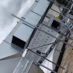 Air System Wentylacje - Montaż kanałów wentylacyjnych na dachu budynku komercyjnego, widok z dołu na szare niebo, elementy konstrukcyjne w trakcie instalacji.