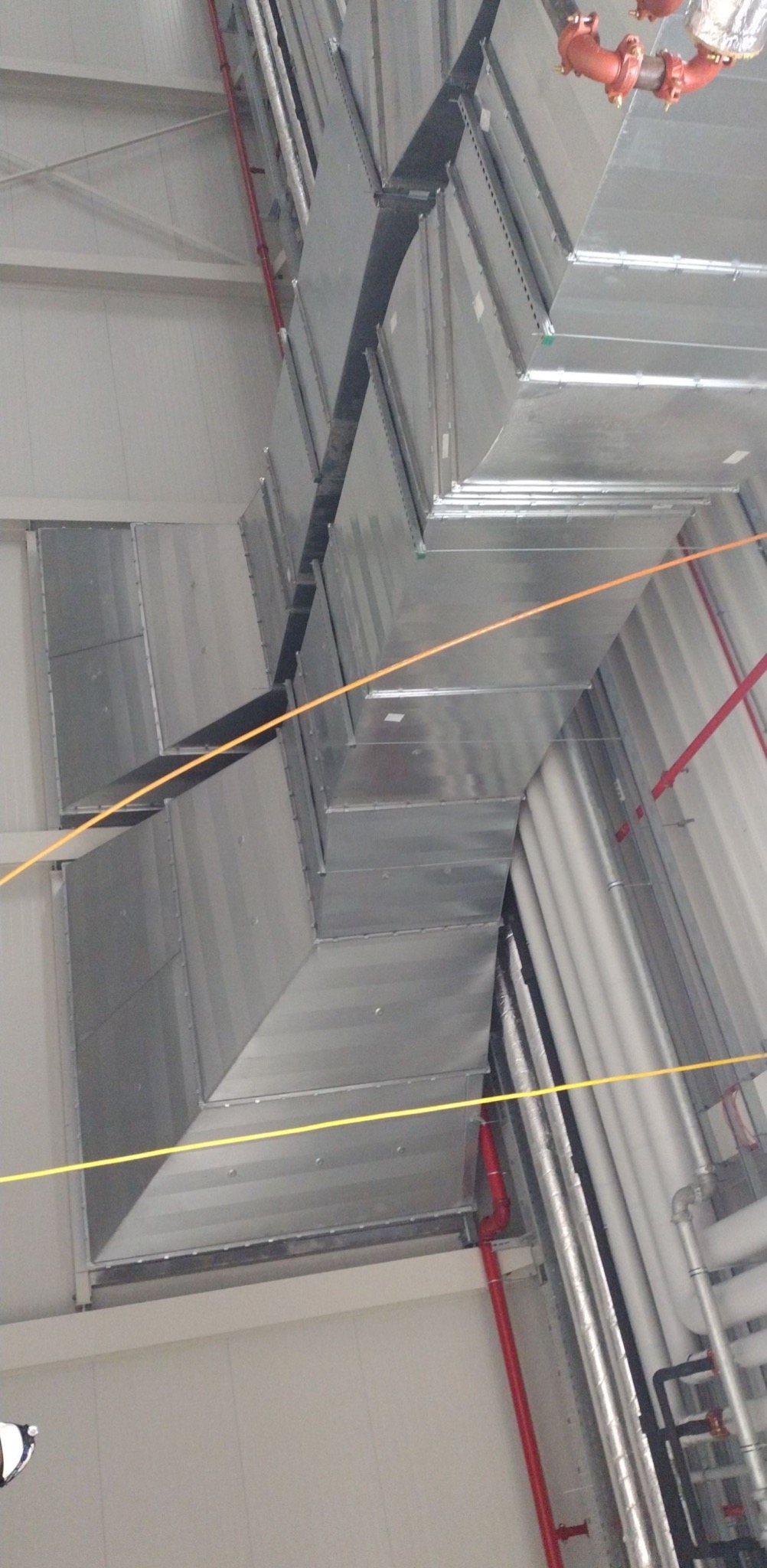 System kanałów wentylacyjnych z blachy ocynkowanej, zamocowany pod sufitem hali przemysłowej, widoczne rury instalacji przeciwpożarowej i inne instalacje.