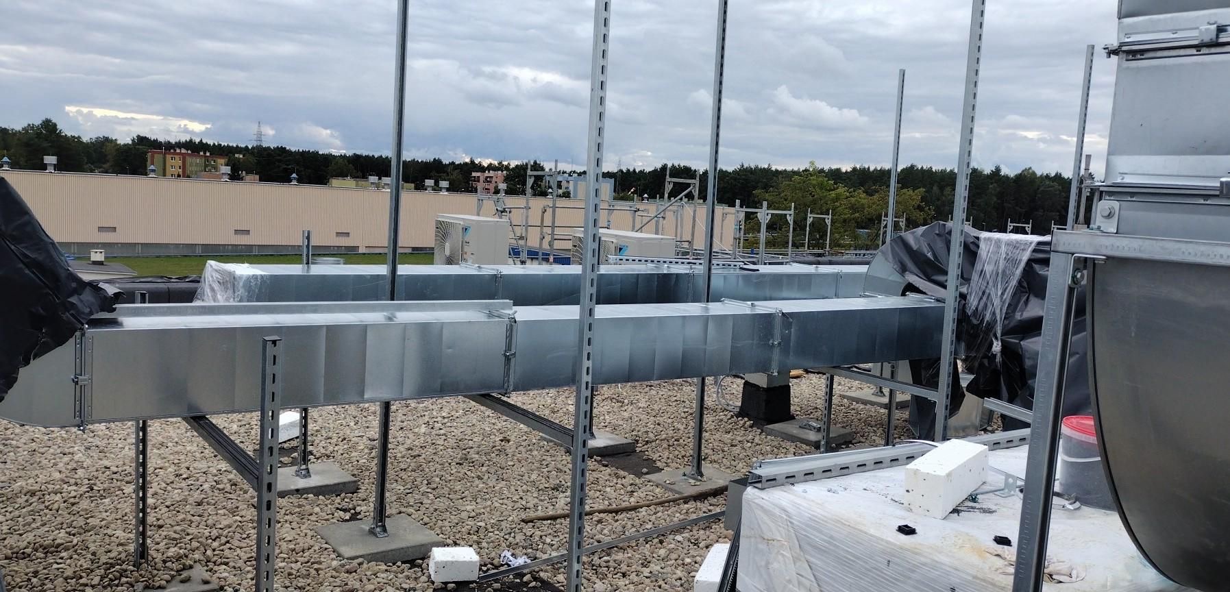 Metalowe kanały wentylacyjne na dachu budynku komercyjnego, wsparte na metalowych konstrukcjach, z widocznymi jednostkami zewnętrznymi klimatyzacji i pokryciem dachu z kamyków.