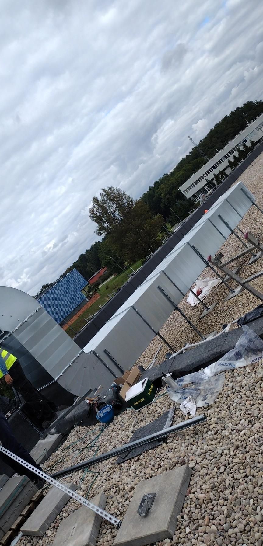 Montaż kanałów wentylacyjnych na dachu budynku komercyjnego, widoczne elementy konstrukcyjne, narzędzia i materiały budowlane, w tle zabudowania i zieleń.