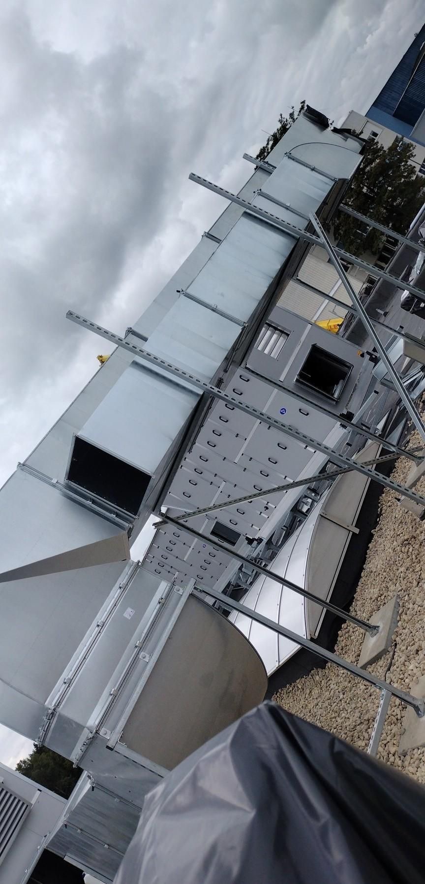 Montaż kanałów wentylacyjnych na dachu budynku komercyjnego, widok z dołu na szare niebo, elementy konstrukcyjne w trakcie instalacji.