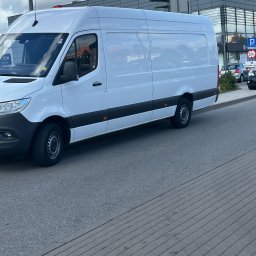 FIRMA HANDLOWO-USŁUGOWA Daniel Wencki - Biały, dostawczy van zaparkowany na ulicy przed budynkiem galerii handlowej w pochmurny dzień.