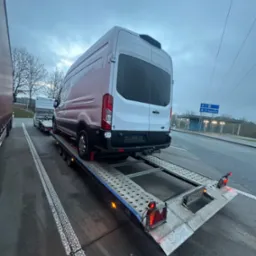 Biały bus na perforowanej platformie lawety, widok z tyłu, tablica rejestracyjna GD, znak drogowy na Regensburg w tle, pochmurne niebo.