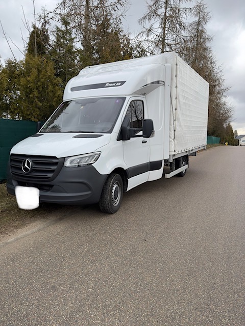 Biały Mercedes-Benz Sprinter z plandeką, zaparkowany na poboczu drogi, widok z boku na tle drzew i pochmurnego nieba.