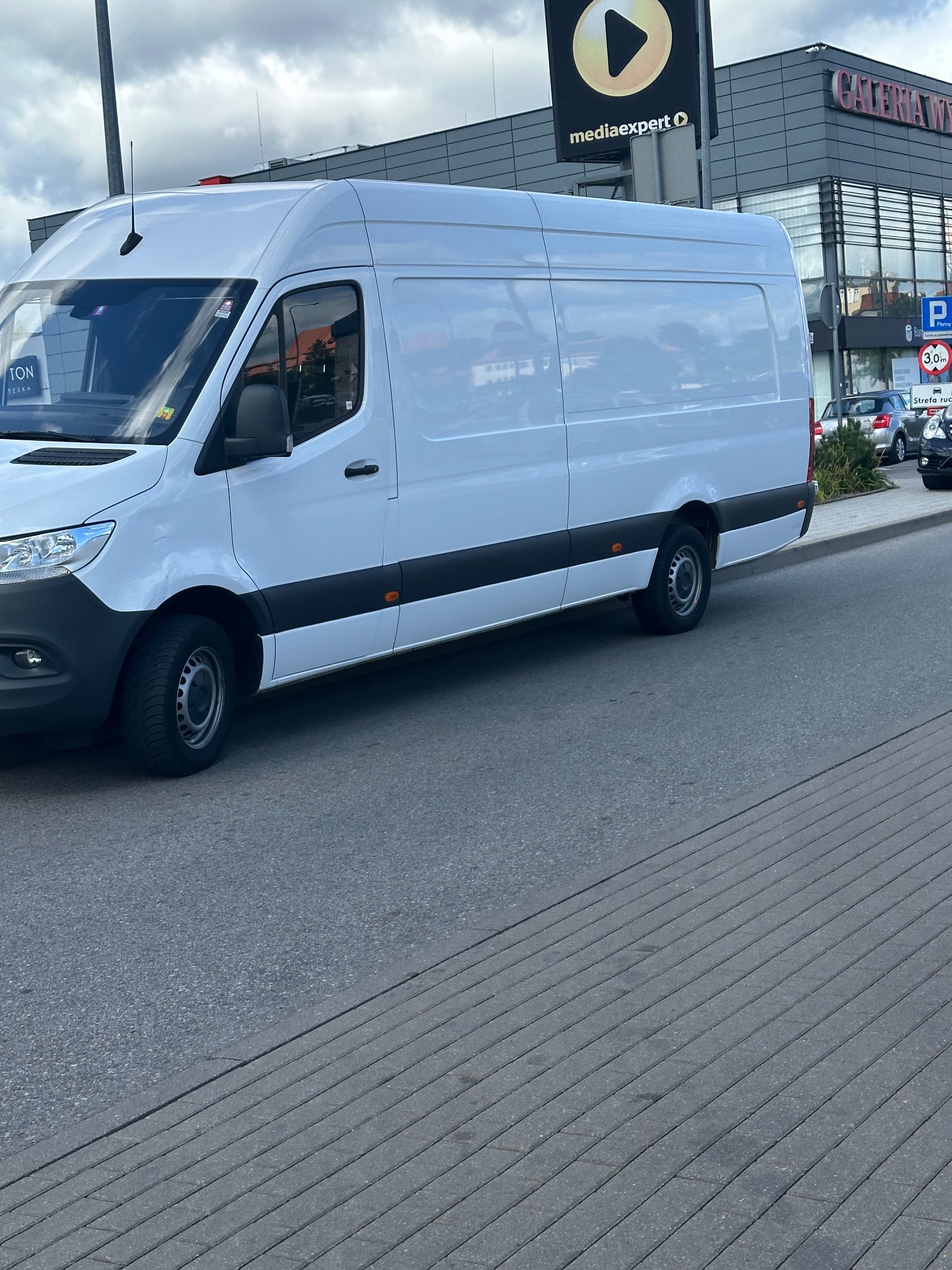 Biały, dostawczy van zaparkowany na ulicy przed budynkiem galerii handlowej w pochmurny dzień.