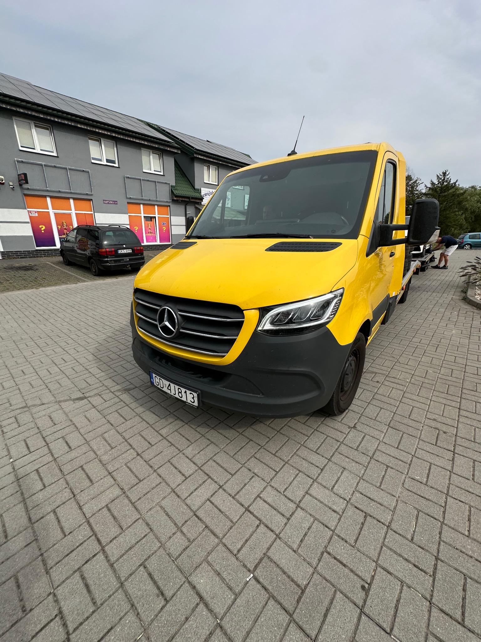 Żółta laweta Mercedes-Benz Sprinter na brukowanym parkingu przed budynkiem z panelami słonecznymi na dachu, w tle zaparkowany czarny samochód osobowy.
