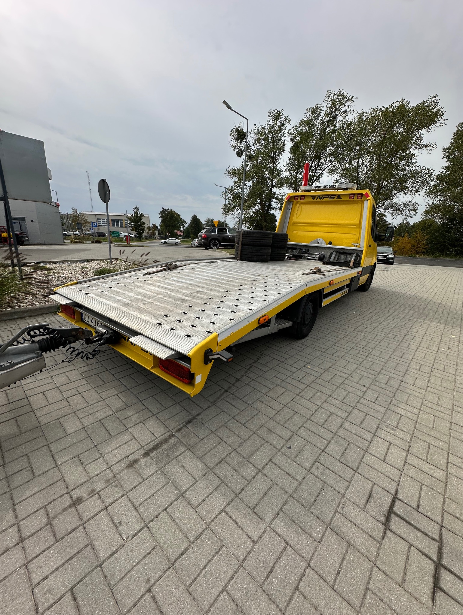 Żółta laweta z platformą załadunkową, na której leżą opony, zaparkowana na brukowanej powierzchni, z widocznym zaczepem holowniczym i elementami otoczenia miejskiego w tle.