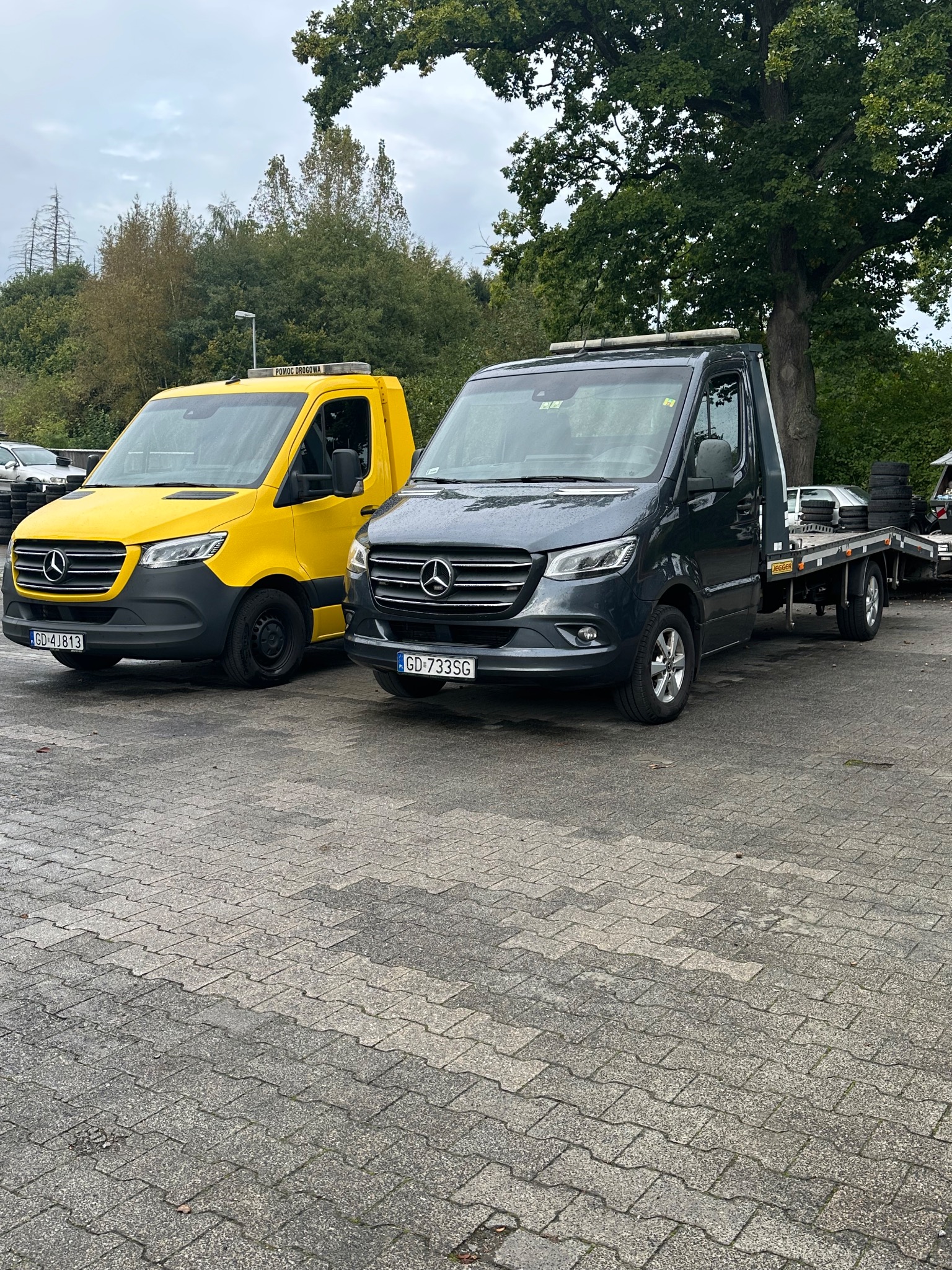 Dwa samochody lawety Mercedes Sprinter: żółty z napisem 'Pomoc Drogowa' i szary, stojące na parkingu z kostki brukowej w pochmurny dzień.