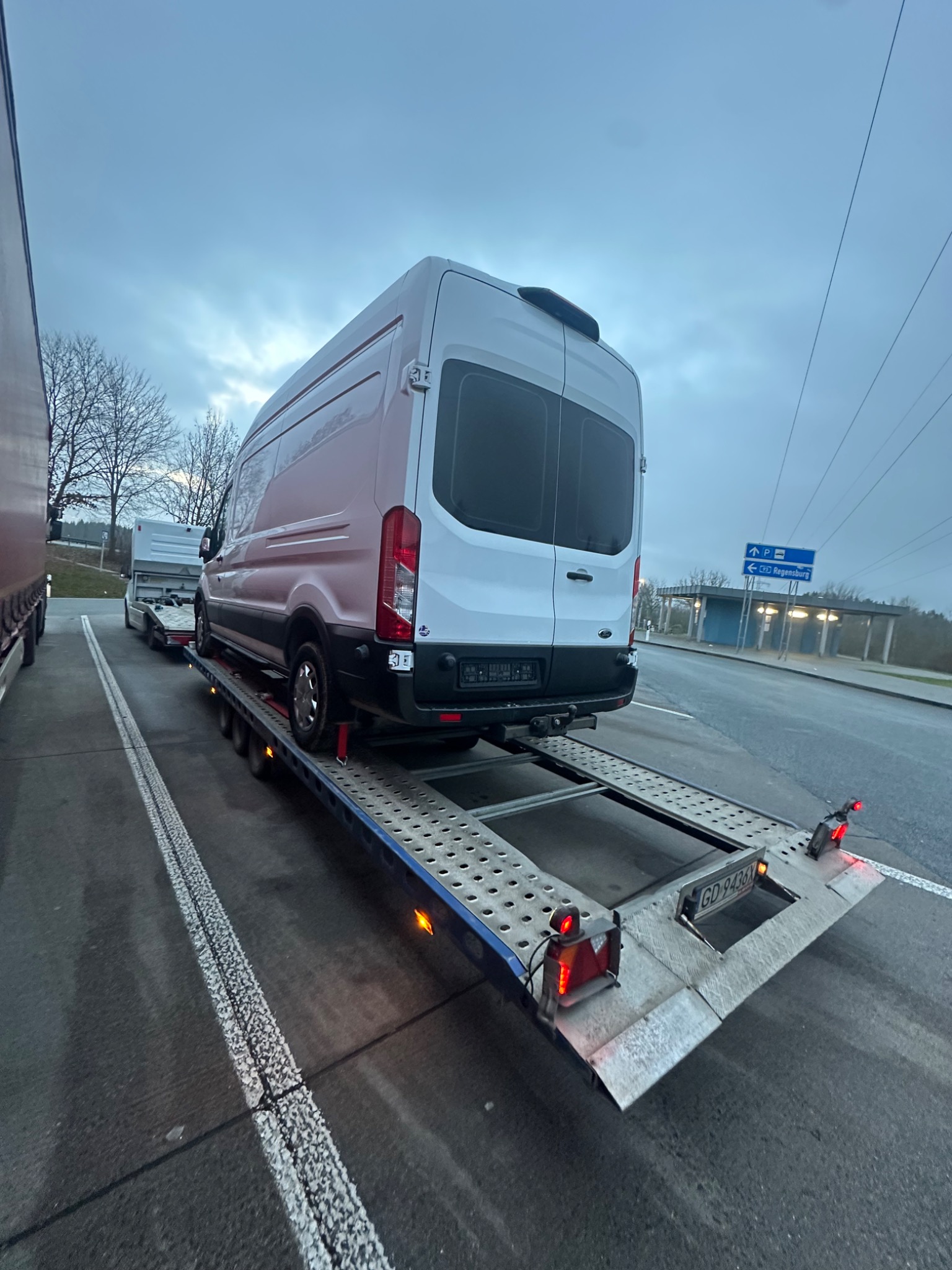 Biały bus na perforowanej platformie lawety, widok z tyłu, tablica rejestracyjna GD, znak drogowy na Regensburg w tle, pochmurne niebo.