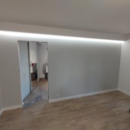 USŁUGI OGÓLNOBUDOWLANE Tomasz Radtke - Nowoczesne wnętrze z przesuwnymi szklanymi drzwiami, podświetlanym sufitem i panelami podłogowymi, widok z perspektywy pokoju z widocznym workiem na podłodze.