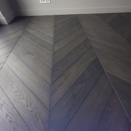 USŁUGI OGÓLNOBUDOWLANE Tomasz Radtke - Ciemne, lakierowane panele ułożone we wzór jodełki francuskiej, widoczne gniazdko elektryczne na ścianie i biała listwa przypodłogowa.