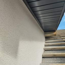 USŁUGI OGÓLNOBUDOWLANE Tomasz Radtke - Fragment elewacji budynku z jasnym, ziarnistym tynkiem, widoczne drewniane elementy konstrukcyjne dachu i fragment niebieskiego nieba w rogu.