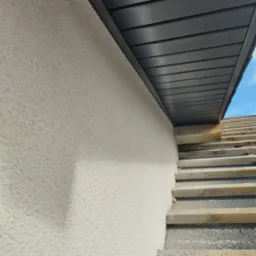 Fragment elewacji budynku z jasnym, ziarnistym tynkiem, widoczne drewniane elementy konstrukcyjne dachu i fragment niebieskiego nieba w rogu.