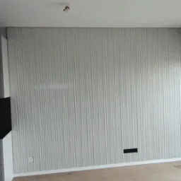 Ściana z szarymi, pionowymi panelami dekoracyjnymi, widoczne gniazdko elektryczne i listwa przypodłogowa, fragment podłogi z jasnego drewna.