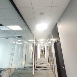 USŁUGI OGÓLNOBUDOWLANE Tomasz Radtke - Korytarz biurowy w trakcie prac wykończeniowych, zabezpieczona podłoga, widoczne szklane ścianki działowe i sufit podwieszany z oświetleniem.