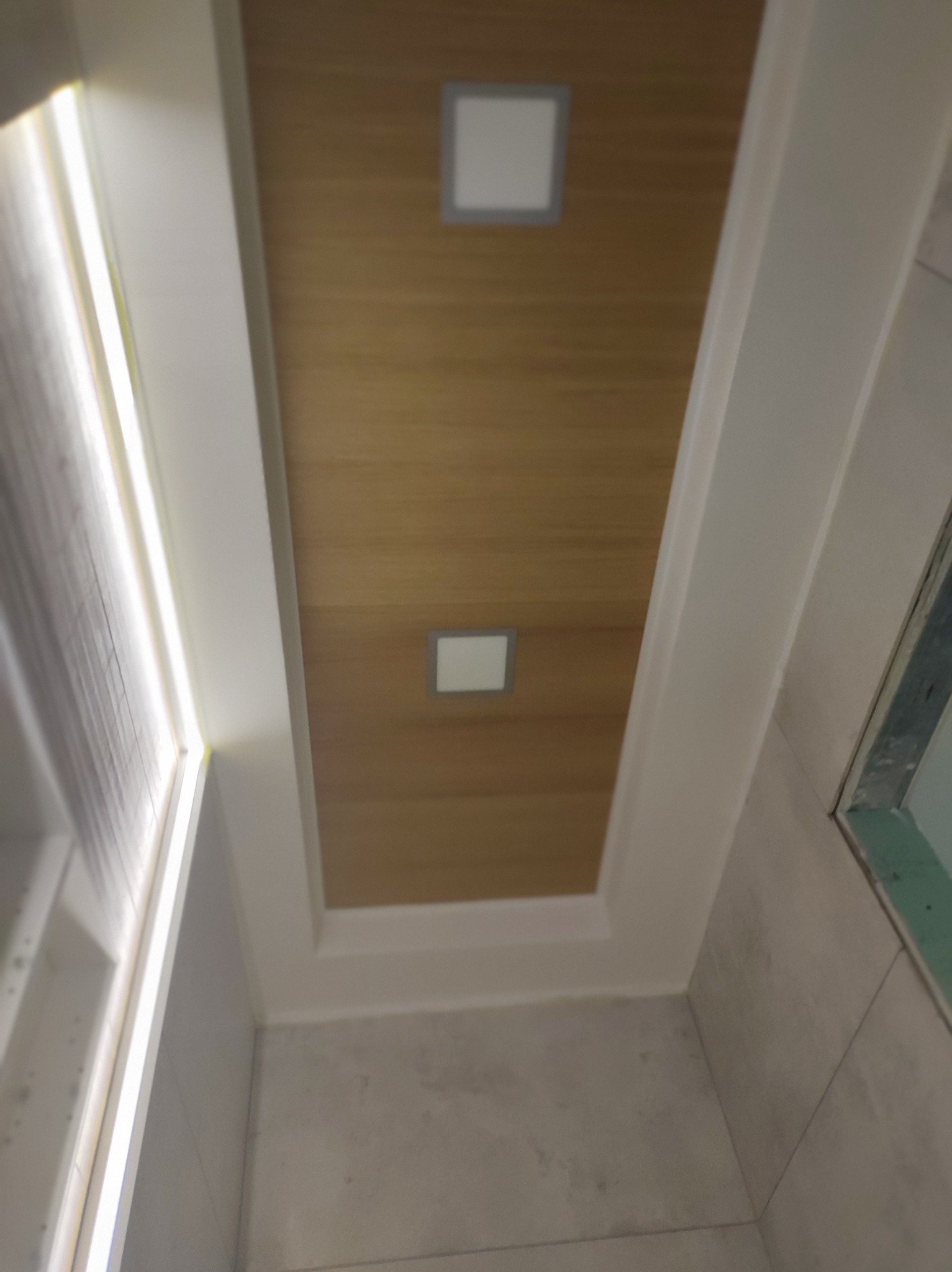 Drewnopodobny sufit z dwoma kwadratowymi lampami wpuszczonymi, obramowany białym, kaskadowym obramowaniem, widok z dołu, fragment ściany z płyt kartonowo-gipsowych po prawej.