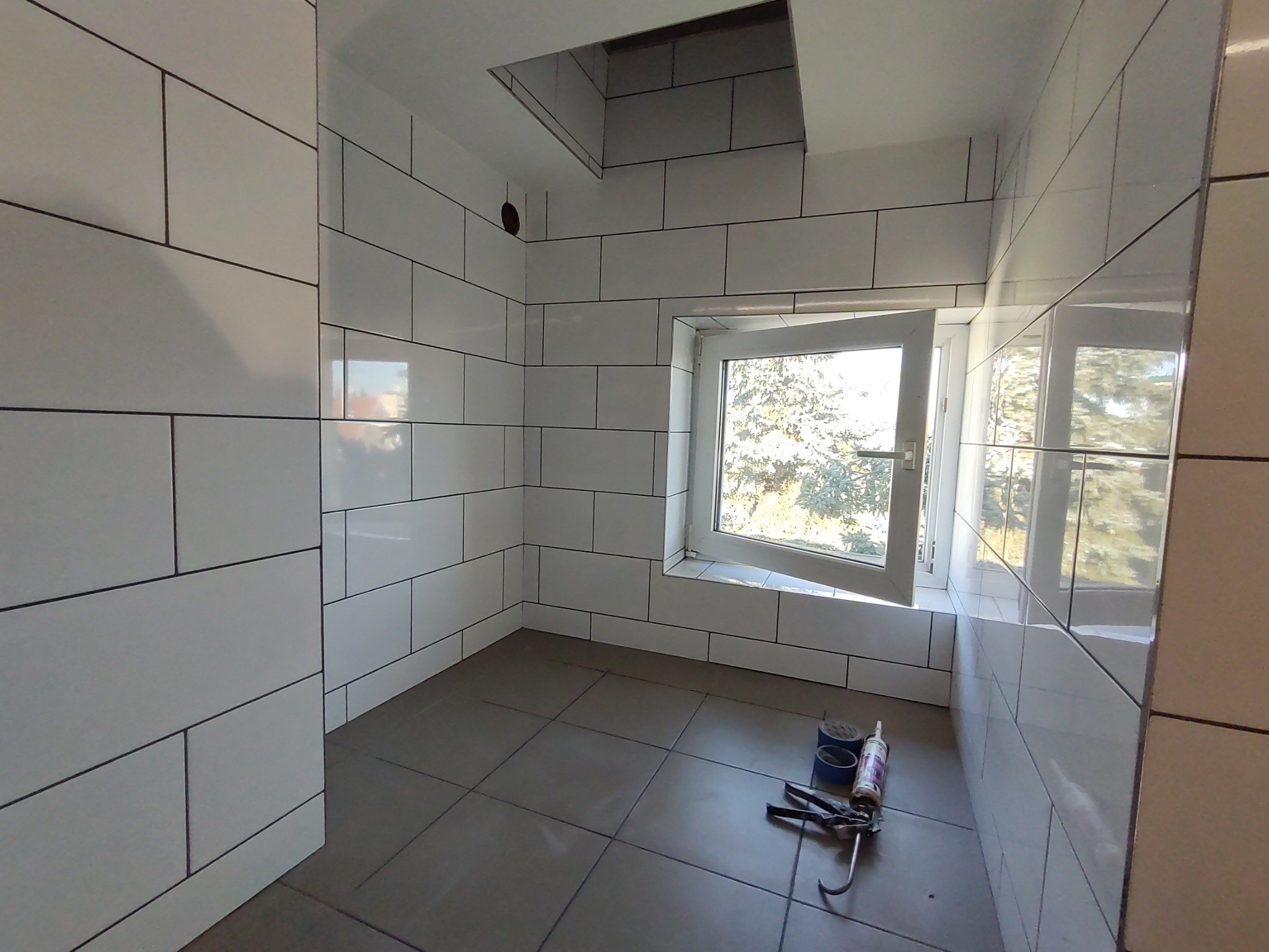 Świeżo wykafelkowana łazienka w Szczecinku z białymi płytkami i szarą podłogą, widoczne narzędzia do fugowania i okno z widokiem na drzewa.