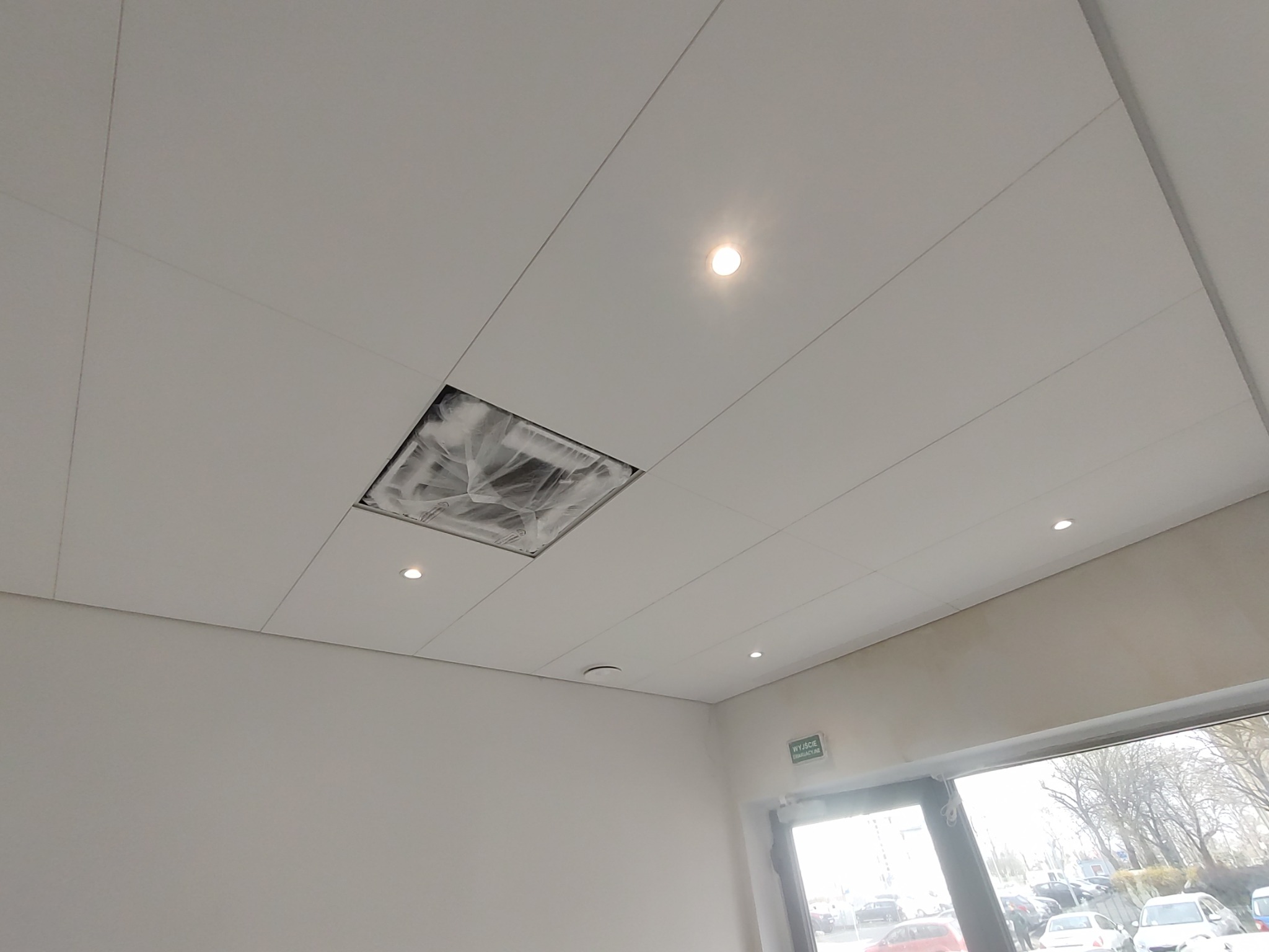 Biały sufit kasetonowy z oświetleniem punktowym, fragment instalacji zakryty folią ochronną, widoczne wyjście ewakuacyjne i parking za oknem.