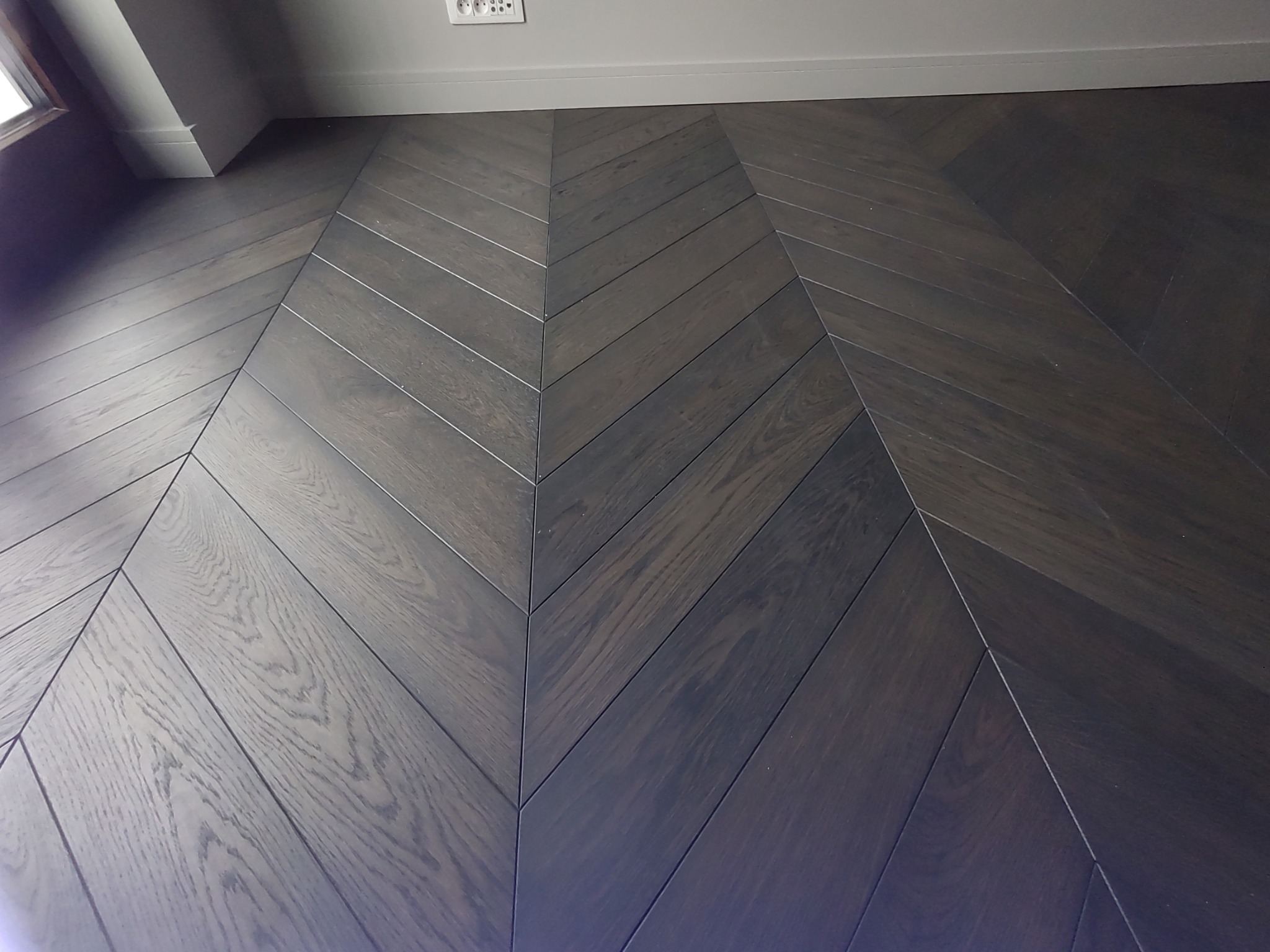 Ciemne, lakierowane panele ułożone we wzór jodełki francuskiej, widoczne gniazdko elektryczne na ścianie i biała listwa przypodłogowa.