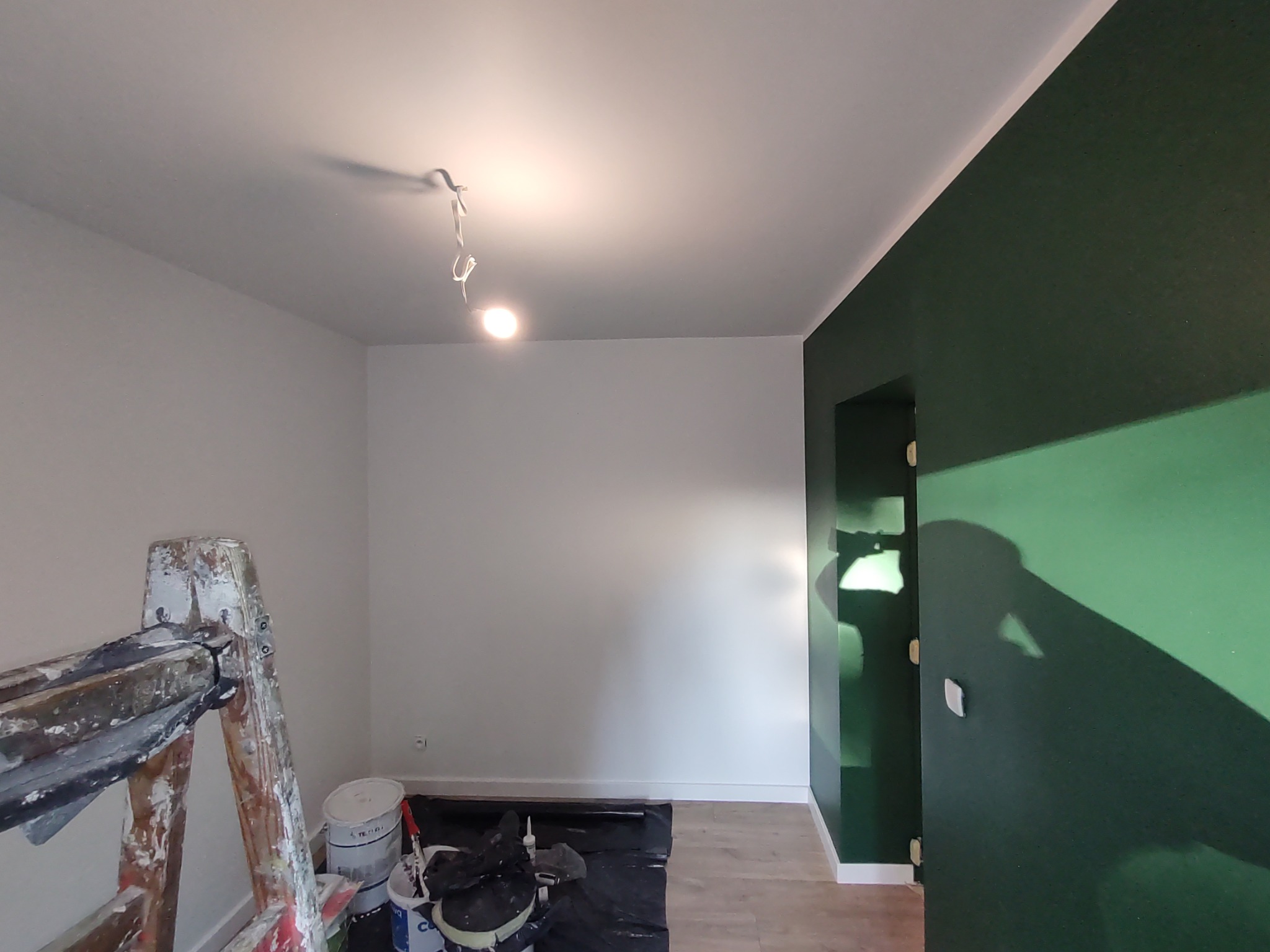 Pokój w trakcie malowania: jedna ściana pomalowana na ciemnozielono, druga biała, widoczna drabina malarska, wiaderka z farbą i folia ochronna na podłodze.