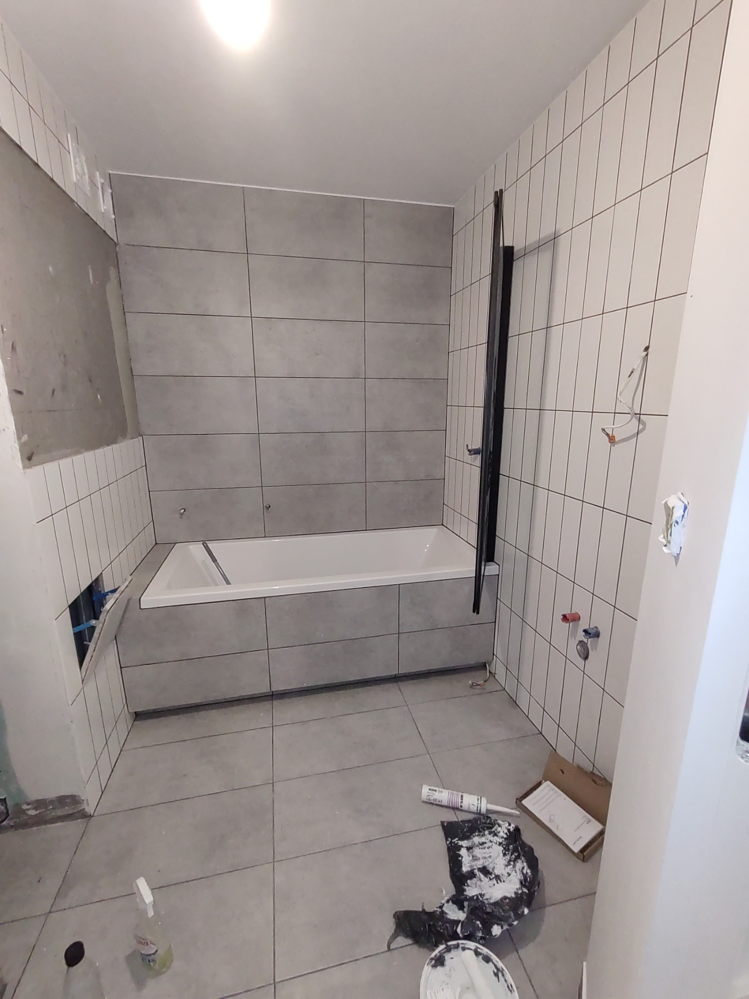 Łazienka w trakcie remontu: częściowo ułożone szare płytki na ścianach, biała wanna obudowana płytkami, widoczne narzędzia i materiały budowlane na podłodze.