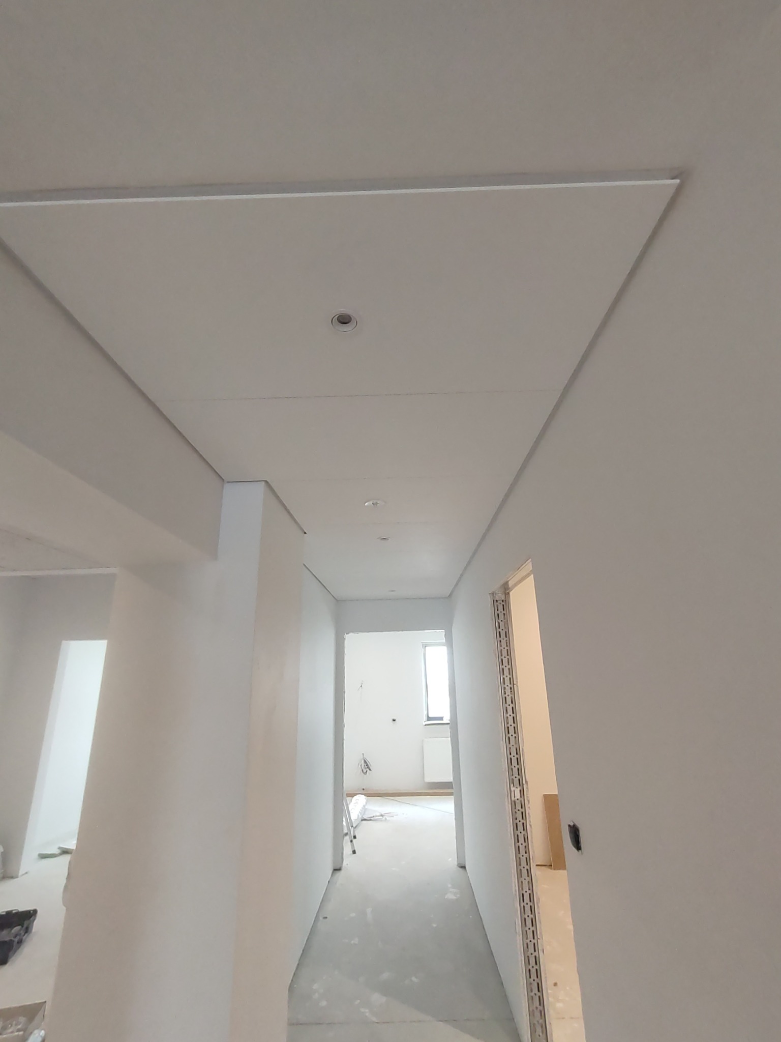 Nowoczesny sufit podwieszany z oświetleniem punktowym w korytarzu, widok z dołu, ściany w trakcie malowania, widoczny fragment drabiny w tle.
