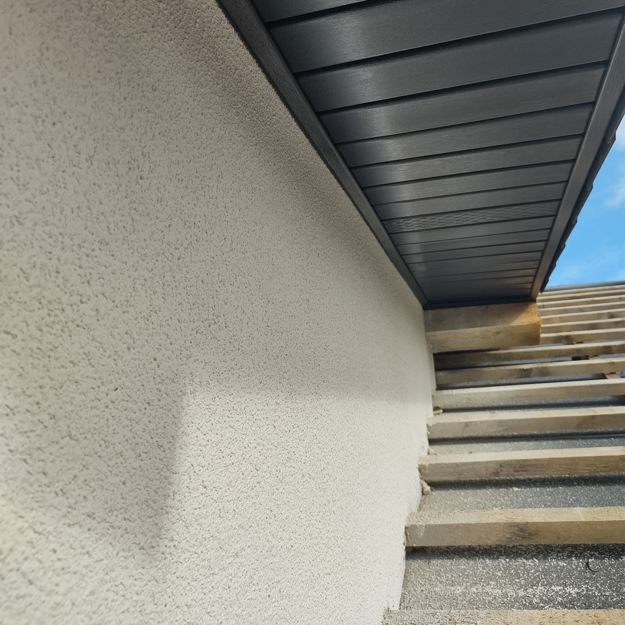 Fragment elewacji budynku z jasnym, ziarnistym tynkiem, widoczne drewniane elementy konstrukcyjne dachu i fragment niebieskiego nieba w rogu.