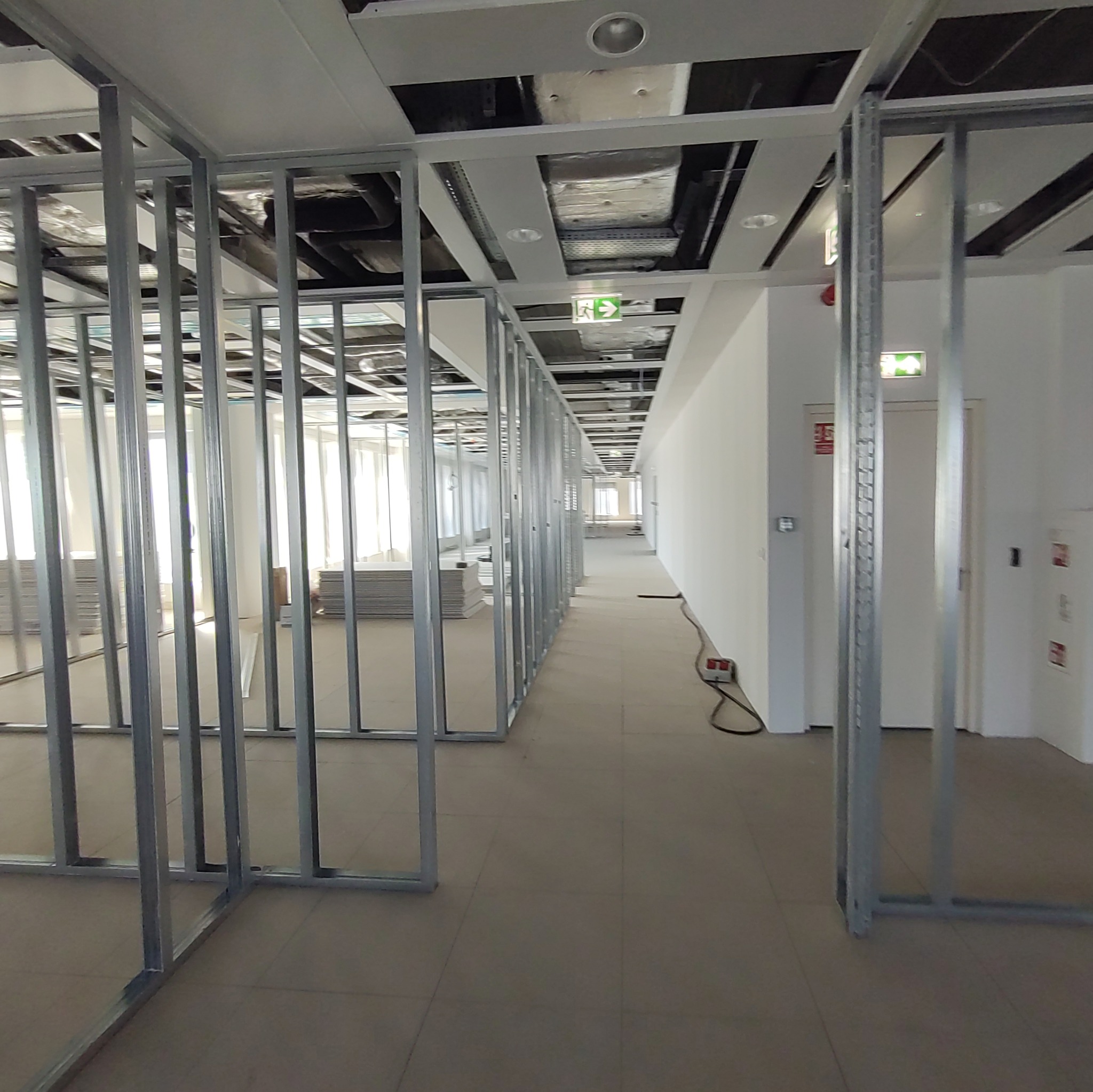 Konstrukcja szkieletowa z profili stalowych wewnątrz budynku biurowego, widoczne płyty gipsowo-kartonowe w tle, instalacje pod sufitem i korytarz z oświetleniem awaryjnym.