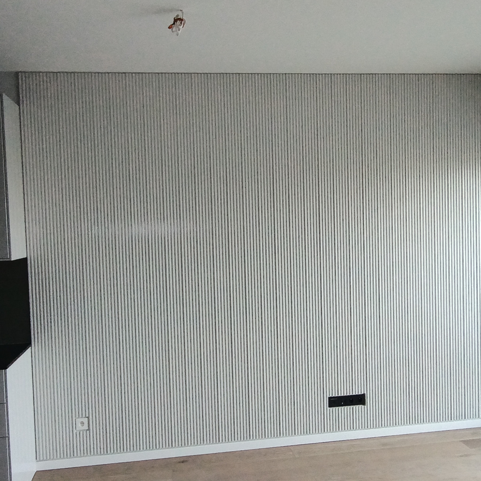 Ściana z szarymi, pionowymi panelami dekoracyjnymi, widoczne gniazdko elektryczne i listwa przypodłogowa, fragment podłogi z jasnego drewna.