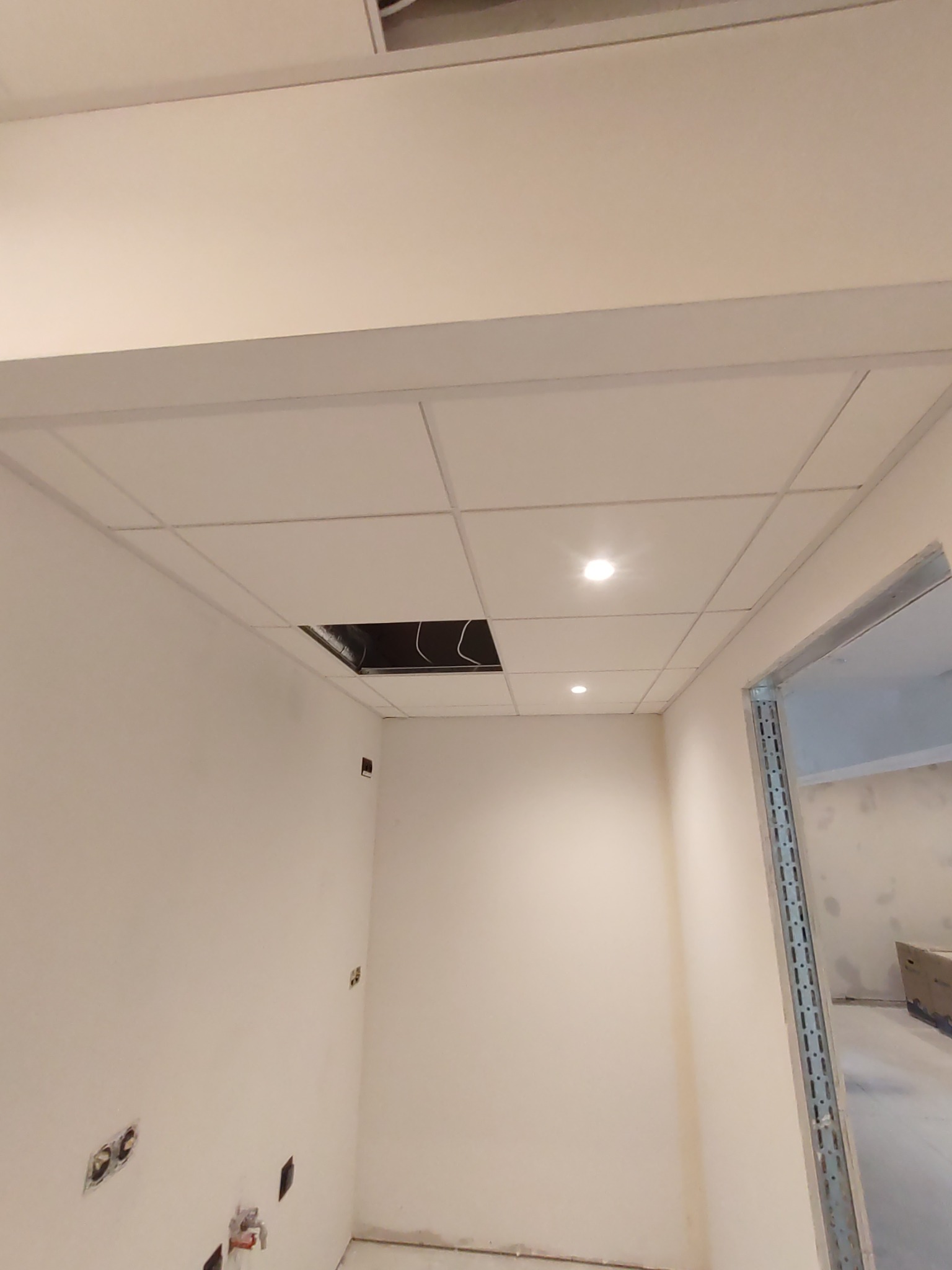 Kasetonowy sufit podwieszany z oświetleniem punktowym i otwartym otworem inspekcyjnym, widoczne niedokończone ściany z gniazdkami elektrycznymi i metalowa ościeżnica drzwiowa.