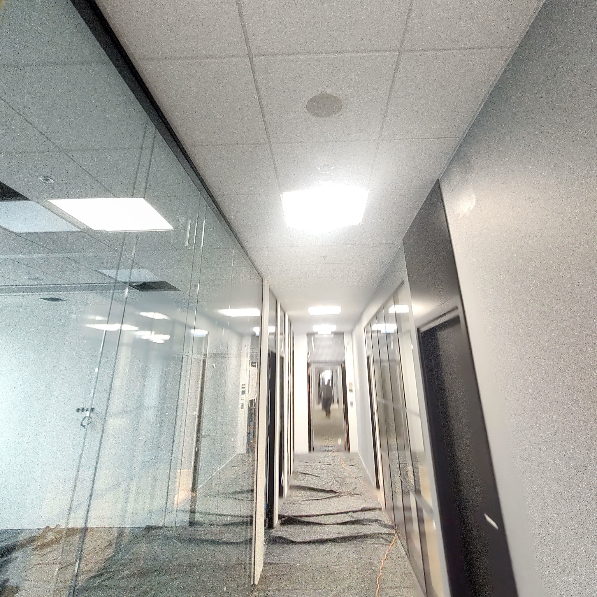 Korytarz biurowy w trakcie prac wykończeniowych, zabezpieczona podłoga, widoczne szklane ścianki działowe i sufit podwieszany z oświetleniem.