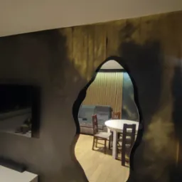 Ściana z efektem dekoracyjnym w odcieniach szarości i złota, widziana z perspektywy salonu w Pleszewie, z nietypowym otworem w kształcie organicznym, ukazującym jadalnię z drewnianym stołem...