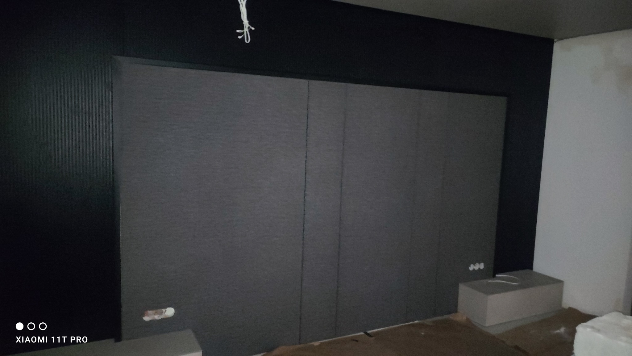 Szara ściana z paneli ściennych z widocznymi listwami dekoracyjnymi, fragment instalacji elektrycznej i niedomalowane fragmenty ściany.