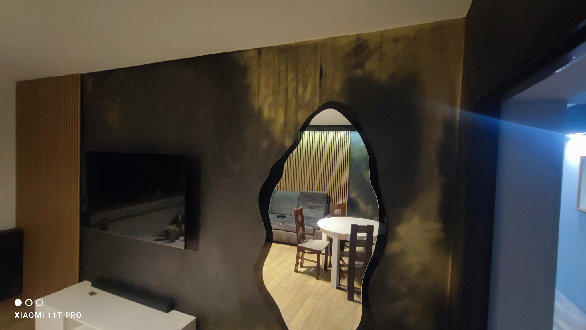 Ściana z efektem dekoracyjnym w odcieniach szarości i złota, widziana z perspektywy salonu w Pleszewie, z nietypowym otworem w kształcie organicznym, ukazującym jadalnię z drewnianym stołem...