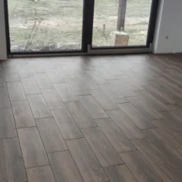 Położona podłoga z płytek imitujących drewno w nowym pomieszczeniu z dużym oknem balkonowym, widoczne niedokończone wykończenia przy ścianach i gniazdkach.