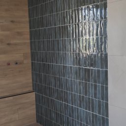 GLAZURNIK PL USŁUGI OGÓLNOBUDOWLANE DAMIAN WIERZBICKI - Wykończona ściana łazienki z ciemnymi, wąskimi płytkami ułożonymi pionowo, kontrastująca z drewnopodobną okładziną i jasną ścianą. Widoczna podłoga z paneli.