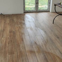 GLAZURNIK PL USŁUGI OGÓLNOBUDOWLANE DAMIAN WIERZBICKI - Jasne, drewnopodobne płytki podłogowe po umyciu, z widocznymi oknami i narzędziami glazurniczymi w tle. Podłoga w trakcie czyszczenia po zakończeniu prac.