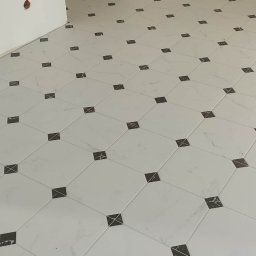 GLAZURNIK PL USŁUGI OGÓLNOBUDOWLANE DAMIAN WIERZBICKI - Biała podłoga z marmurową fakturą i czarnymi, kwadratowymi wstawkami w nowym wnętrzu. Widoczny fragment ściany z gniazdkiem i przejście do kolejnego pomieszczenia.