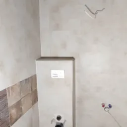 Wnętrze łazienki w trakcie wykańczania: widok na stelaż podtynkowy WC z przyłączami, częściowo ułożone płytki ścienne i podłogowe imitujące drewno.