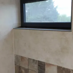 Wykończona łazienka z oknem: beżowe płytki na ścianach, dekoracyjny pas z brązowych płytek, czarna rama okna z widokiem na zieleń. Nowoczesne i estetyczne rozwiązanie.