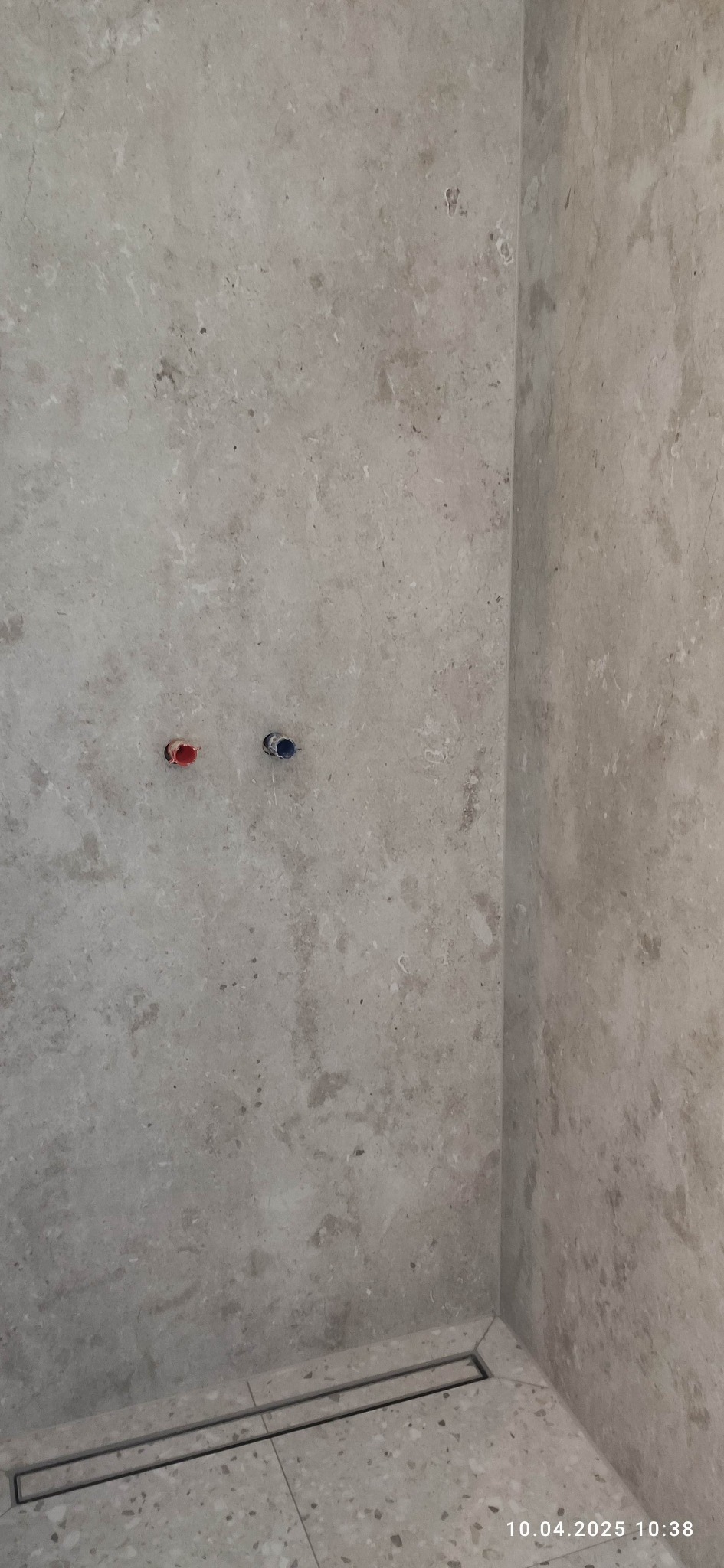 Wykończona ściana i podłoga w łazience z odpływem liniowym. Widoczne przyłącza wody ciepłej i zimnej. Płytki imitujące kamień w odcieniach szarości.